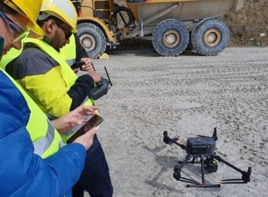 Two workers in protective clothing and helmets operate a drone on a construction site using a remote control and a smartphone, with a construction vehicle in the background.