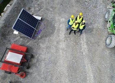 Drone shot of three construction workers in safety vests and helmets on a construction site, next to a solar panel, a machine, and a vehicle.