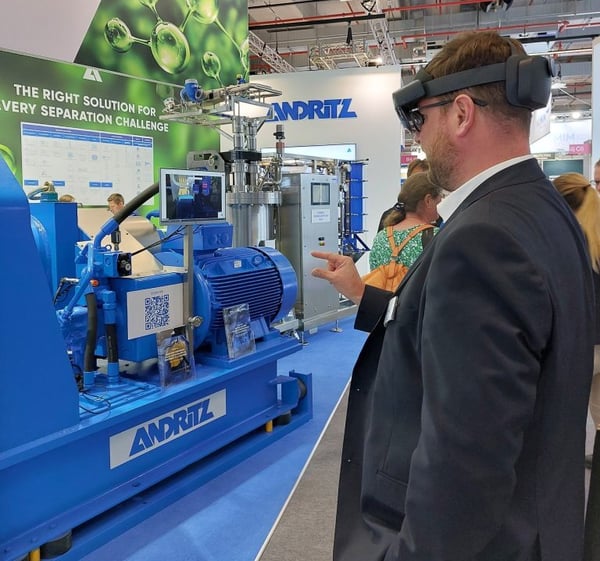 Man with AR glasses observes a large blue Andritz industrial plant at a trade fair; additional machine data is displayed on a screen.