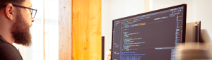 Bearded programmer working at a computer screen displaying code in a development environment.